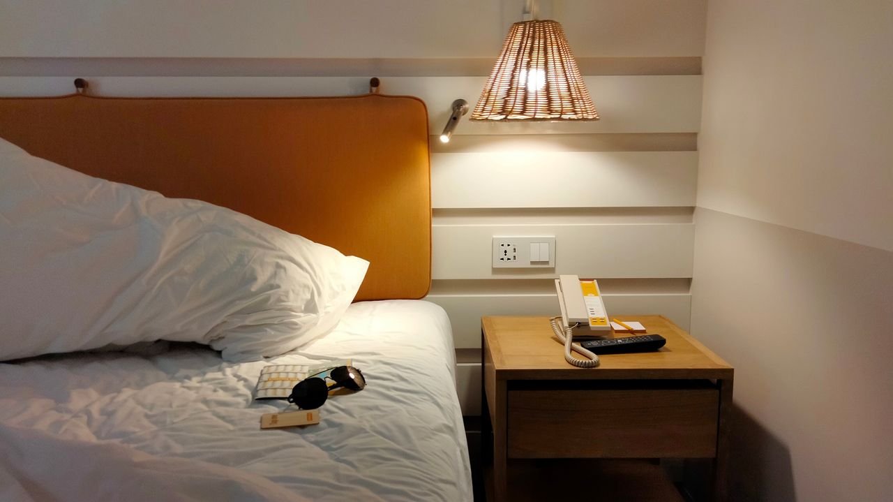 Mastering the First Impression: Your intriguing post title goes here Inviting bedroom space with a lamp-lit nightstand, phone, and sunglasses on the bed.